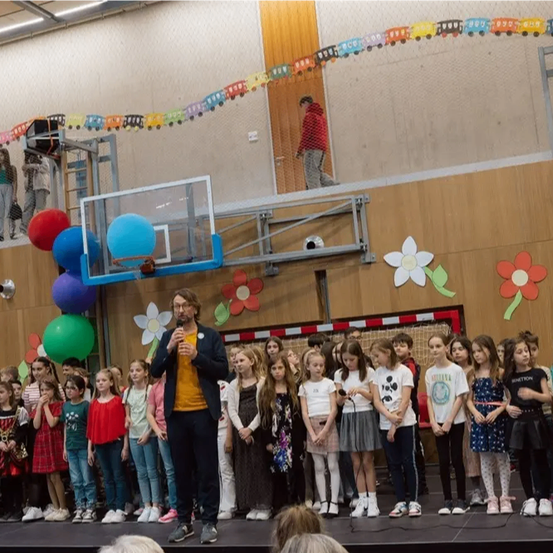 Ein Mann steht mit einem Mikrofon auf der Bühne und spricht eine Reihe von Kindern an. Hinter ihm sind bunte Luftballons an einem Basketballkorb befestigt, und eine Person läuft an der Decke. Die Wand ist mit Blumen und Luftballons dekoriert.