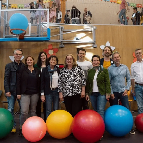 Eine Gruppe Erwachsener posiert für ein Foto in einem Raum voller großer bunter Übungsbälle. Die Wände sind mit Blumen und Luftballons dekoriert. Ein Basketballkorb ist an der Wand montiert.