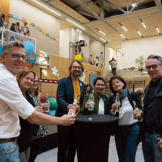 Eine Gruppe von Menschen steht um einen Tisch und hält Flaschen. Sie scheinen eine Feier zu veranstalten. Einige Luftballons sind an den Geländern befestigt, und Menschen gehen im Hintergrund.
