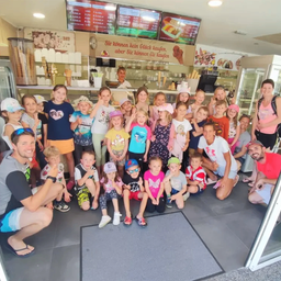 Eine Gruppe von Kindern und Erwachsenen posiert für ein Foto in einem Eiscafé, lächelt und macht verschiedene Handgesten.