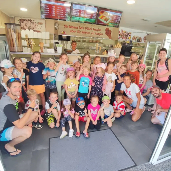 Eine Gruppe von Kindern und Erwachsenen posiert für ein Foto in einem Eiscafé, lächelt und macht verschiedene Handgesten.