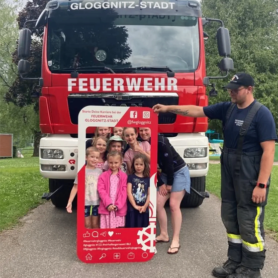 Ein Feuerwehrmann in Uniform steht mit einem Fotorahmen vor einem Feuerwehrauto und hält Kinder.