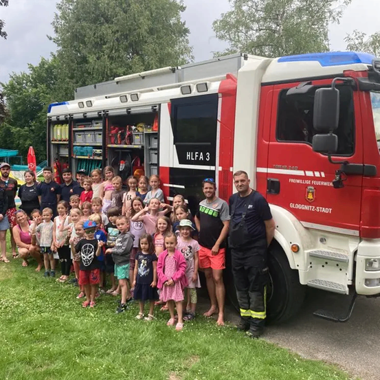 Eine Gruppe von Kindern und Erwachsenen steht neben einem roten Feuerwehrauto. Das Auto ist mit HLFA 3 gekennzeichnet.