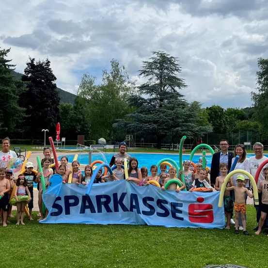 Eine Gruppe von Kindern und Erwachsenen posiert für ein Foto am Pool mit einem Banner, das 'SPARKASSE' steht, unter einem bewölkten Himmel.