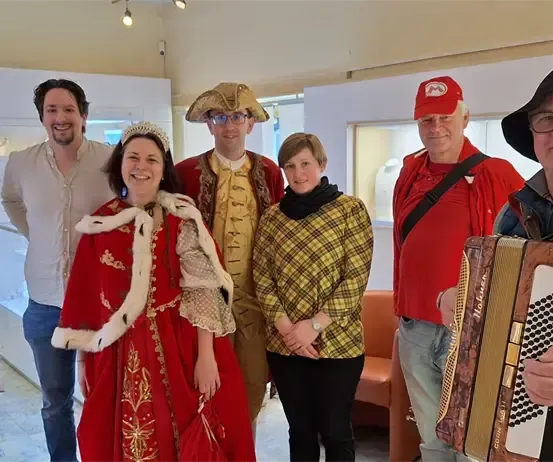 Eine Gruppe von Menschen in verschiedenen Kostümen, darunter eine Frau in einem roten Kleid, ein Mann mit Piratenhut und ein Mann, der Akkordeon spielt, posiert für ein Foto in einem Raum.