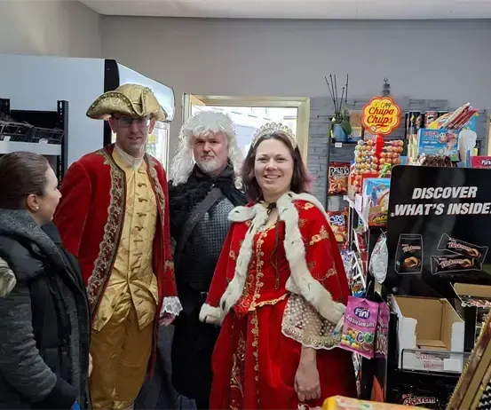 Drei Personen in historischen Kostümen stehen in einem Geschäft. Eine Frau in einem roten Kleid und Krone lächelt. Ein Mann in einem roten und goldenen Kostüm steht neben ihr. Ein weiterer Mann mit Perücke und Brille schaut zu. Verschiedene Produkte sind auf Regalen und Tischen ausgestellt.