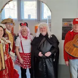 Eine Gruppe von Menschen in verschiedenen Kostümen posiert für ein Foto in einem Innenbereich mit einem großen Bogentor. Sie halten Musikinstrumente. Hinter ihnen befindet sich eine Wand mit einer Tafel.