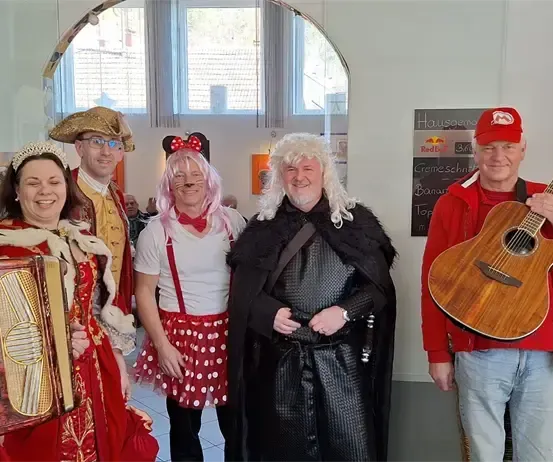 Eine Gruppe von Menschen in verschiedenen Kostümen posiert für ein Foto in einem Innenbereich mit einem großen Bogentor. Sie halten Musikinstrumente. Hinter ihnen befindet sich eine Wand mit einer Tafel.