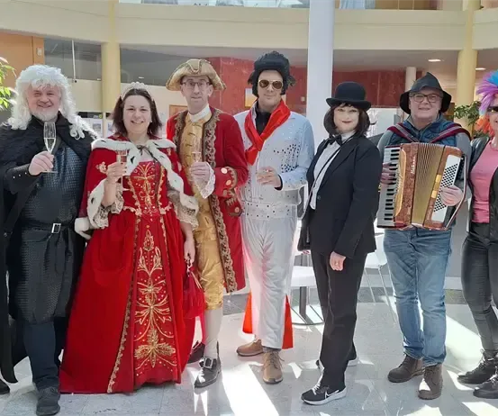 Eine Gruppe von Menschen in verschiedenen Kostümen, mit Wein glasses in der Hand, posiert für ein Foto in einem Saal mit weißem Boden und Topfpflanzen.