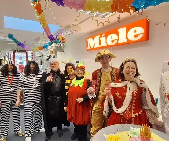 Gruppe von Menschen in verschiedenen Kostümen posiert für ein Foto vor einem Tisch mit Kuchen, Getränken und einem Miele-Schild an der Wand.