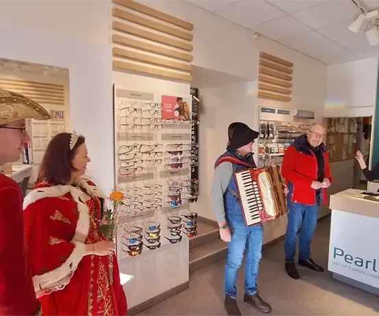 Eine Gruppe von Menschen in Kostümen befindet sich in einem Geschäft. Ein Akkordeonspieler steht unter ihnen, während andere ein Gespräch führen. Das Geschäft verfügt über eine Glasvitrine mit einer Auswahl an Brillen und einem Schild mit der Aufschrift 'Pearle.'
