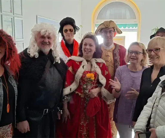 Eine Gruppe von Menschen in Kostümen posiert für ein Foto in einem Raum. Eine Frau in einem roten Kleid mit einer Blume hält eine Kamera.