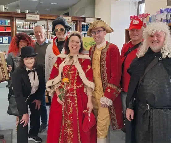 Eine Gruppe von Menschen in Kostümen, einer hält ein Akkordeon, steht in einem Raum mit Regalen und Gegenständen im Hintergrund.