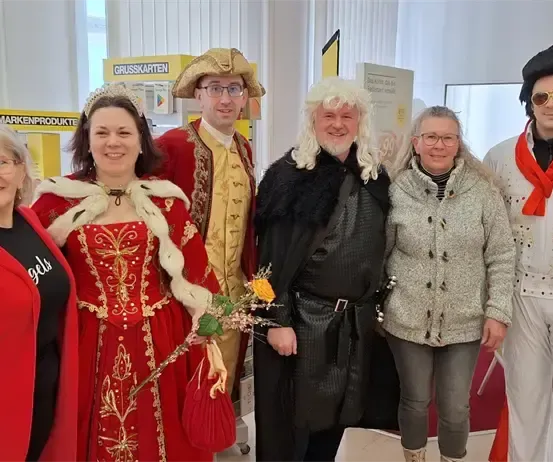 Sechs Menschen posieren für ein Foto in einem Raum mit gelber Wand und Schildern. Eine Person ist in einem roten Kostüm gekleidet, eine andere in einem weißen Elvis-Kostüm und andere in verschiedenen Outfits.