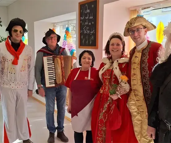 Eine Gruppe von Menschen in Kostümen posiert für ein Foto, darunter eine Frau mit Blumen, ein Mann mit einem Akkordeon und andere in verschiedenen historischen Kostümen gekleidet.