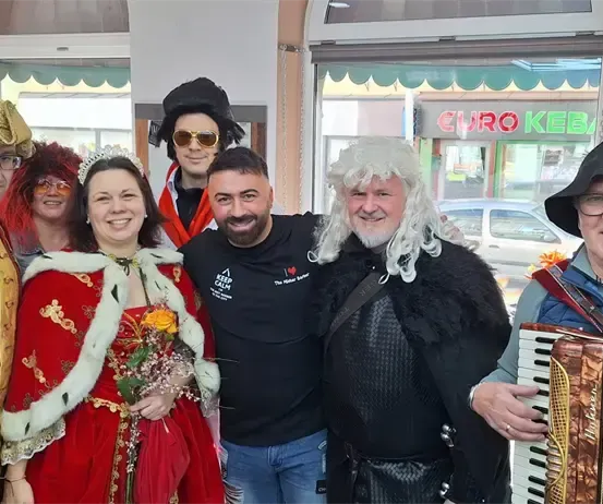 Eine Gruppe von Menschen in verschiedenen Kostümen, darunter ein Ritter, eine Königin und ein Mann mit einem Akkordeon, posiert für ein Foto vor einem Geschäft.