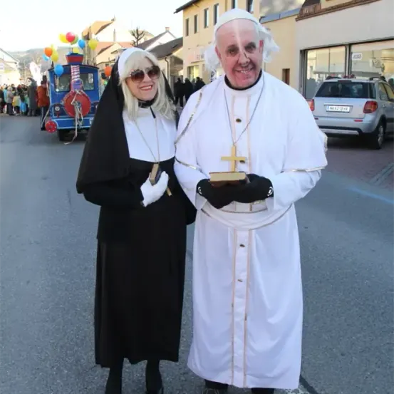 Zwei Menschen, verkleidet als Nonne und Papst, stehen auf einer Straße. Hinter ihnen sind Gebäude, ein Auto und eine Parade mit Luftballons.