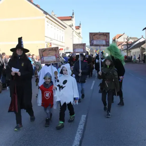 Eine Gruppe von Menschen in Kostümen geht die Straße entlang, mit Gebäuden und einer Kirche im Hintergrund. Einige halten Schilder hoch, andere tragen Körbe.