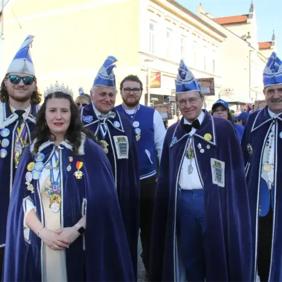 Eine Gruppe von Menschen in traditioneller Kleidung, möglicherweise für ein Fest, posiert für ein Foto. Eine Frau mit Krone steht in der Mitte, während andere um sie herum Hüte tragen und Medaillen an ihren Roben haben. Hinter ihnen sind Gebäude und Straßenlaternen zu sehen.