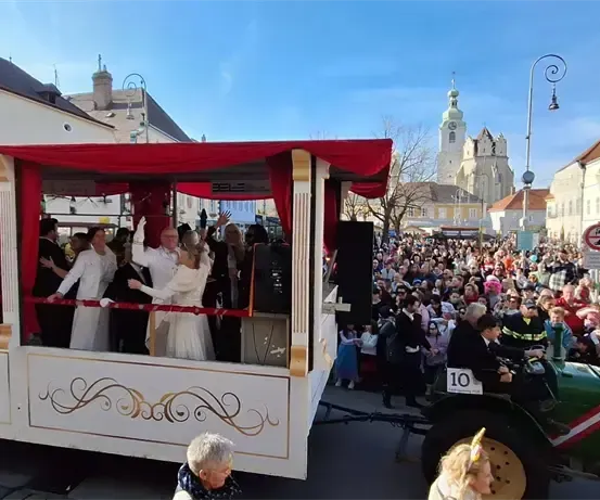 Ein festlicher Umzug mit Menschen in weißen und roten Gewändern, die auf einem geschmückten Wagen reiten, der von einem Traktor gezogen wird, vor einer großen Menschenmenge.