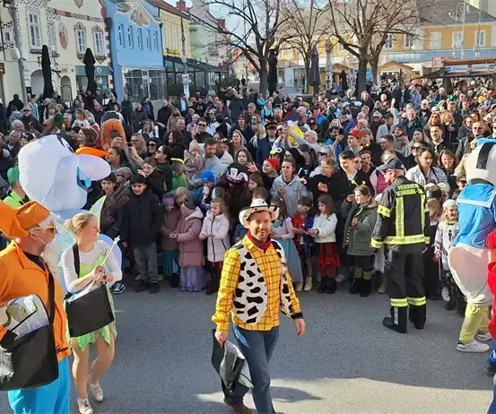 Eine große Menschenmenge in Kostümen und Masken versammelt sich auf einer Straße, mit Maskottchen und Gebäuden im Hintergrund.