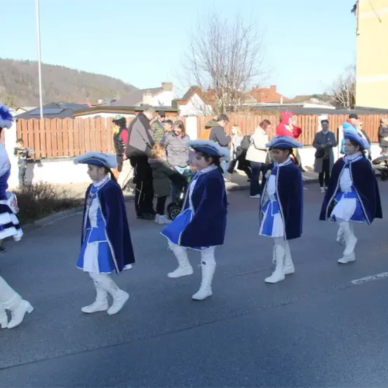 Eine Gruppe von Kindern in blauen Kostümen und Mützen läuft bei einem Umzug, begleitet von Zuschauern an einem sonnigen Tag.