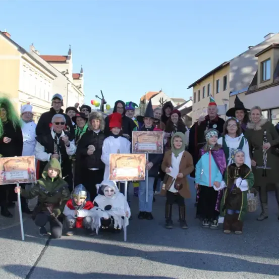 Eine Gruppe von Menschen in Kostümen, einige mit Schildern, steht in einer Reihe auf einer Stadtstraße. Sie sind von Gebäuden umgeben, und einige tragen Hüte und Brillen. Einige halten Musikinstrumente.