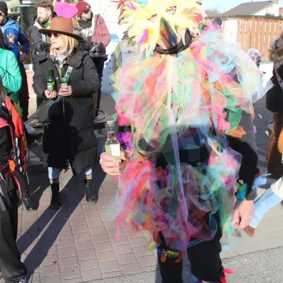 Eine Person in einem farbenfrohen gefiederten Kostüm hält eine Flasche in der Hand und geht auf einer Straße, mit anderen um sie herum, einschließlich einer Frau in einer Mütze, die zwei Flaschen hält.