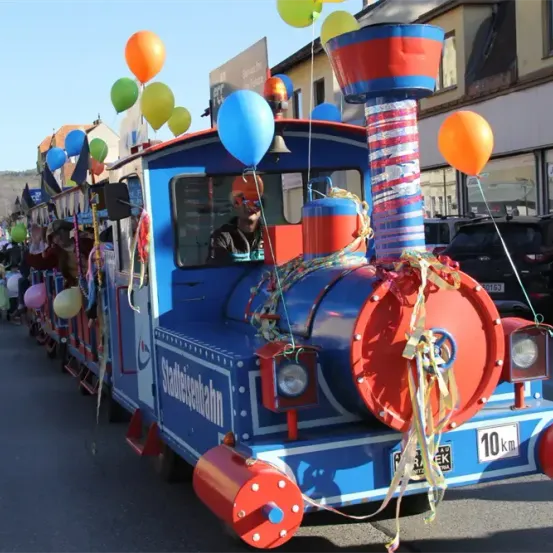 Ein geschmückter Zug mit einem Fahrer, der einen orangefarbenen Turban und eine Sonnenbrille trägt, führt einen Umzug an, mit bunten Luftballons und Bändern, an einem sonnigen Tag.