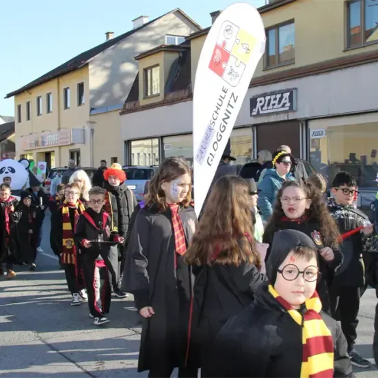 Eine Gruppe von Kindern in verschiedenen Kostümen, darunter einige in Hogwarts-Roben, zieht bei einem Umzug die Straße entlang. Im Hintergrund befinden sich Gebäude und ein Banner mit der Aufschrift Gloggnitz.
