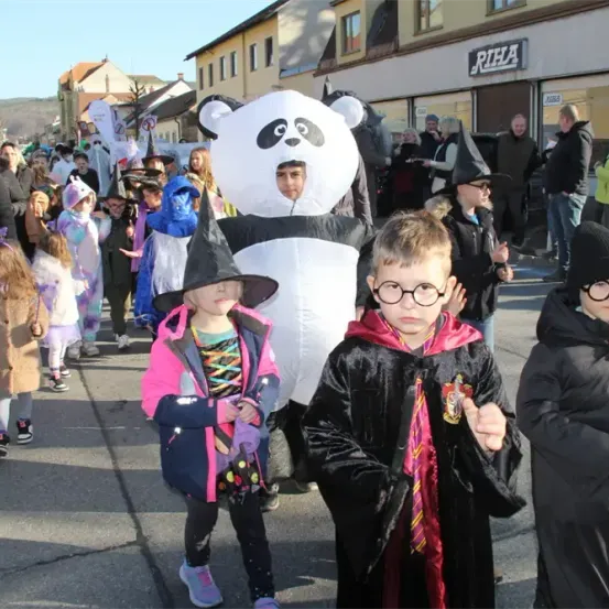 Eine Gruppe von Kindern in Kostümen geht in einem Umzug mit einer Person in einem Panda-Kostüm. In der Nähe steht eine Menge Menschen und schaut zu.