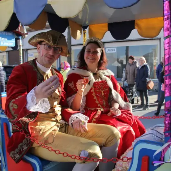Ein Mann und eine Frau in königlichen Kostümen sitzen in einer bunten Kutsche, lächeln, mit einer Menschenmenge im Hintergrund.