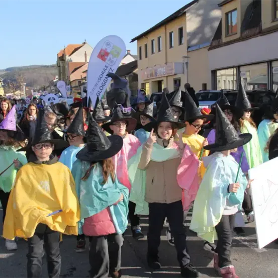 Eine Gruppe von Kindern in Hexenkostümen läuft in einem Umzug. Einige tragen Brillen und Hüte, andere halten Schilder. Gebäude und geparkte Autos sind im Hintergrund.