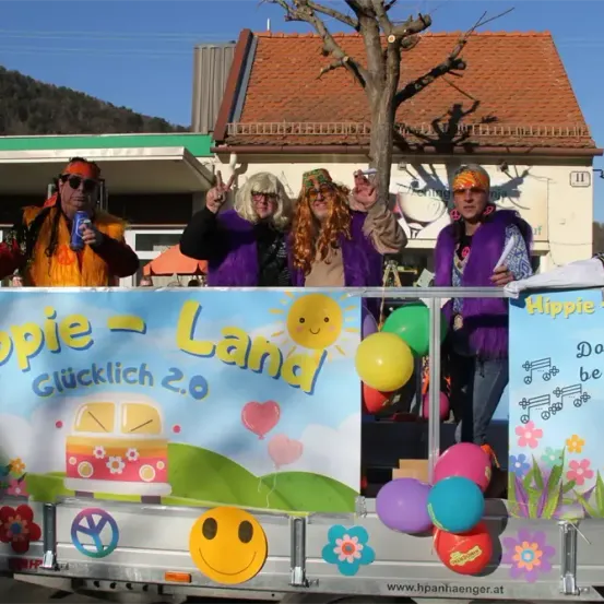 Eine Gruppe von Menschen steht auf einem dekorierten Truck, gekleidet in Hippie-Kostümen, mit einem Banner, das 'Hippie Land Glücklich 2.0' und verschiedene Ballons zeigt. Sie stehen vor einem Gebäude mit einem roten Dach.