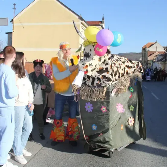 Eine Person in einem Kuhkostüm mit Luftballons geht auf der Straße mit Zuschauern, einer mit einem Mikrofon, vor Gebäuden und einem klaren Himmel.