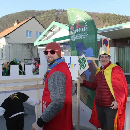 Zwei Erwachsene in roten Kostümen mit Kronen stehen neben einem Zaun mit einer grünen Fahne. Eine Person trägt einen roten Hut und eine Sonnenbrille. Ein Haus und ein Berg sind im Hintergrund.