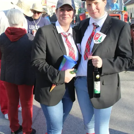 Zwei Frauen in Schuluniformen und Mützen, eine hält eine Flasche und die andere einen blauen Becher, stehen in einer Menschenmenge mit einem Feuerwehrwagen im Hintergrund.