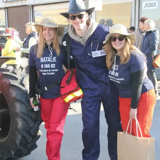 Drei Personen posieren vor einem großen Reifen. Eine Frau hält eine Flasche, eine andere hat eine Handtasche, und ein Mann in einem blauen Uniform mit einem Stethoskop lächelt.