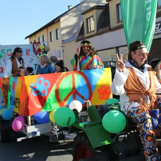 Eine Gruppe von Menschen in farbenfrohen Kostümen fährt mit einem geschmückten Traktor bei einer Parade. Sie halten ein Banner mit einem Friedenszeichen.