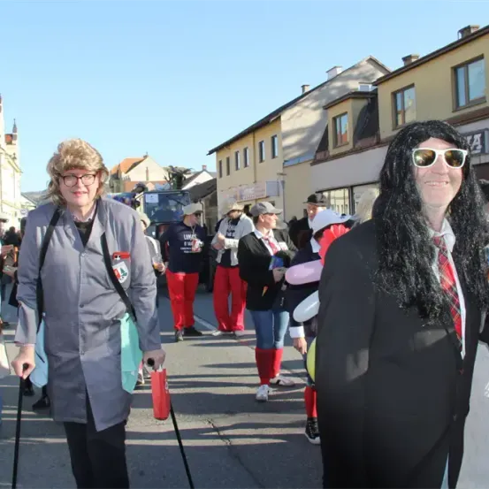 Zwei Erwachsene in Kostümen gehen während einer Parade auf einer Straße mit mehreren anderen Menschen. Gebäude säumen die Straße, einige mit Schildern.
