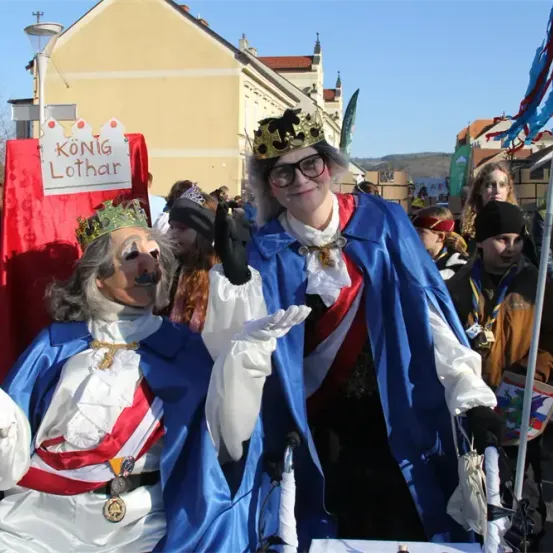 Zwei Personen, die als Monarchen gekleidet sind, tragen Kronen und Kostüme und stehen bei einer Parade, wobei eine Person ein rotes Schild mit der Aufschrift 'KoniG Lothar' hält. Hinter ihnen nehmen mehrere Personen an der Parade teil, und ein gelbes Gebäude ist im Hintergrund zu sehen.