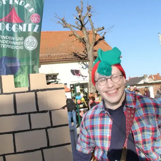 Ein Mann in einem bunten karierten Hemd und einer grünen Elfenmütze mit einer Quaste steht vor einer Mauer aus Pappsteinen und lächelt. Hinter ihm befindet sich ein Gebäude mit einem roten Dach, das ein Banner mit der Aufschrift 'finder und nderinnnen terreichs' trägt. Ein blattloser Baum ist zu sehen, und im Hintergrund gehen Menschen umher.