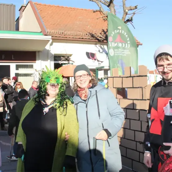 Drei Menschen in Kostümen stehen vor einem Gebäude. Die Frau trägt eine grüne Drachenmaske. Hinter ihnen hält ein Mann mit Brille eine Ziegelmauer.