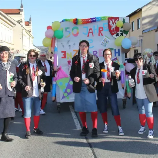 Eine Gruppe von Menschen, die Schuluniformen und rote Socken tragen, posiert für ein Foto vor einem Wagen, auf dem '50 Jahre' steht. Sie halten verschiedene Gegenstände wie Becher, Flaschen und Stöcke. Dahinter wird ein Banner mit bunten Luftballons und einem Logo gezeigt.