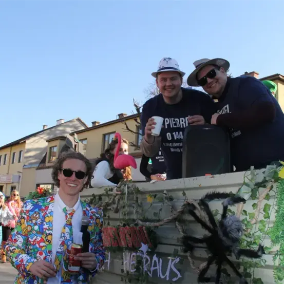 Drei Männer in bunten Kostümen, einer in einem Anzug, stehen hinter einem geschmückten Wagen mit einer Spinne. Der Mann in der Mitte hält einen Becher, während die anderen in die Kamera schauen. Dahinter eine Frau mit einem rosa Flamingo und Gebäuden im Hintergrund.