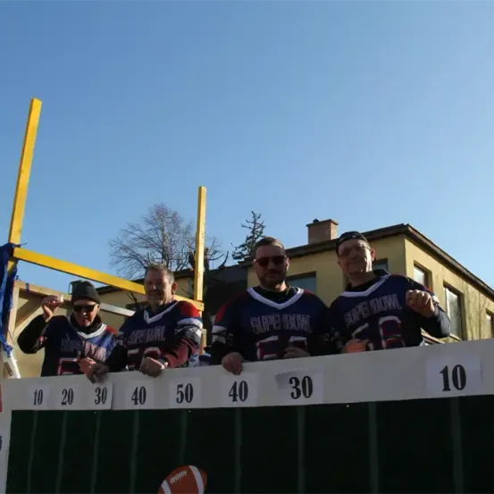 Vier Männer in Super Bowl Trikots stehen vor einer Anzeigetafel mit den Zahlen 10, 20, 30, 40, 50 und 30. Sie posieren für ein Foto. Dahinter steht ein Haus mit Bäumen.