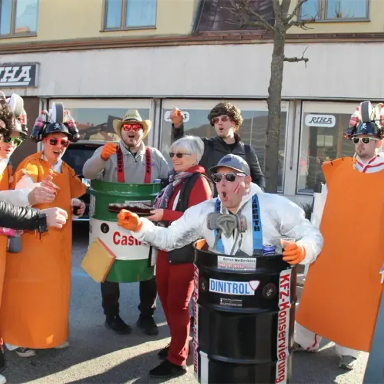 Eine Gruppe von Menschen in orangefarbenen Kostümen, einer in einem weißen Anzug, steht vor einem Gebäude mit dem Wort 'Riha'. Sie halten einen grünen und schwarzen Fass mit 'Casu' darauf.