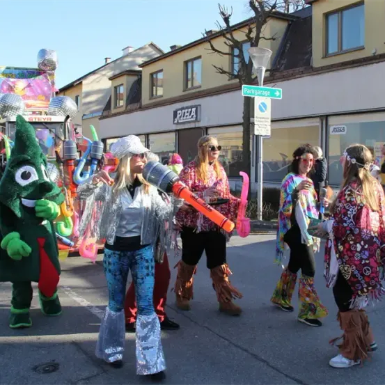 Eine Gruppe von Menschen in leuchtenden Kostümen und Stiefeln zieht durch die Straße, eine Person in einem großen grünen Kostüm mit einem Regenschirm, eine andere hält ein großes orangefarbenes Objekt.