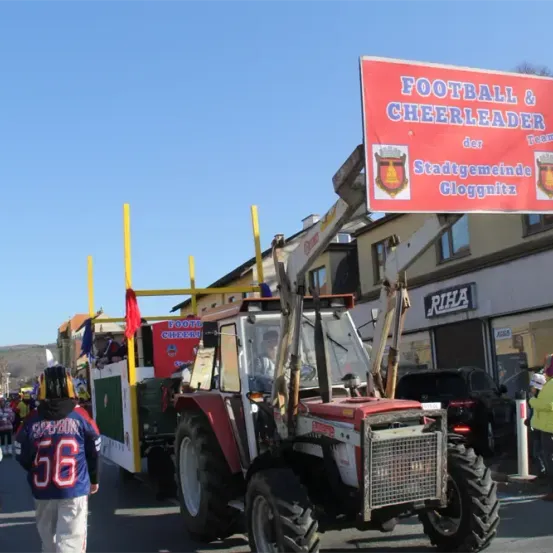 Ein roter Traktor, dekoriert mit einem Fußball- und Cheerleader-Thema, führt eine Parade an, wobei eine Menge Menschen zuschaut.