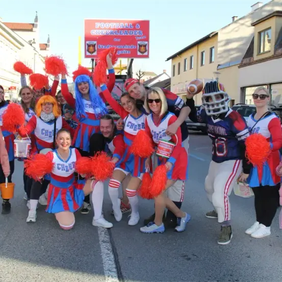 Eine Gruppe von Football-Cheerleadern posiert für ein Foto auf der Straße, mit einem Schild für Football und Cheerleader über ihnen. Sie tragen rote, weiße und blaue Uniformen.